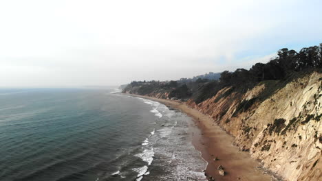 drone aéreo disparado sobre las olas azules del océano pacífico frente a los acantilados de la playa en la costa nublada de santa bárbara, california