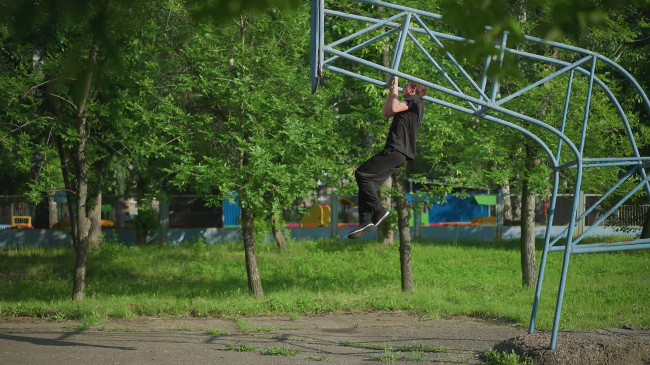 A young boy dressed in a black outfit gradually performs pull-ups on outdoor equipment, set against a backdrop of lush green trees and a fence