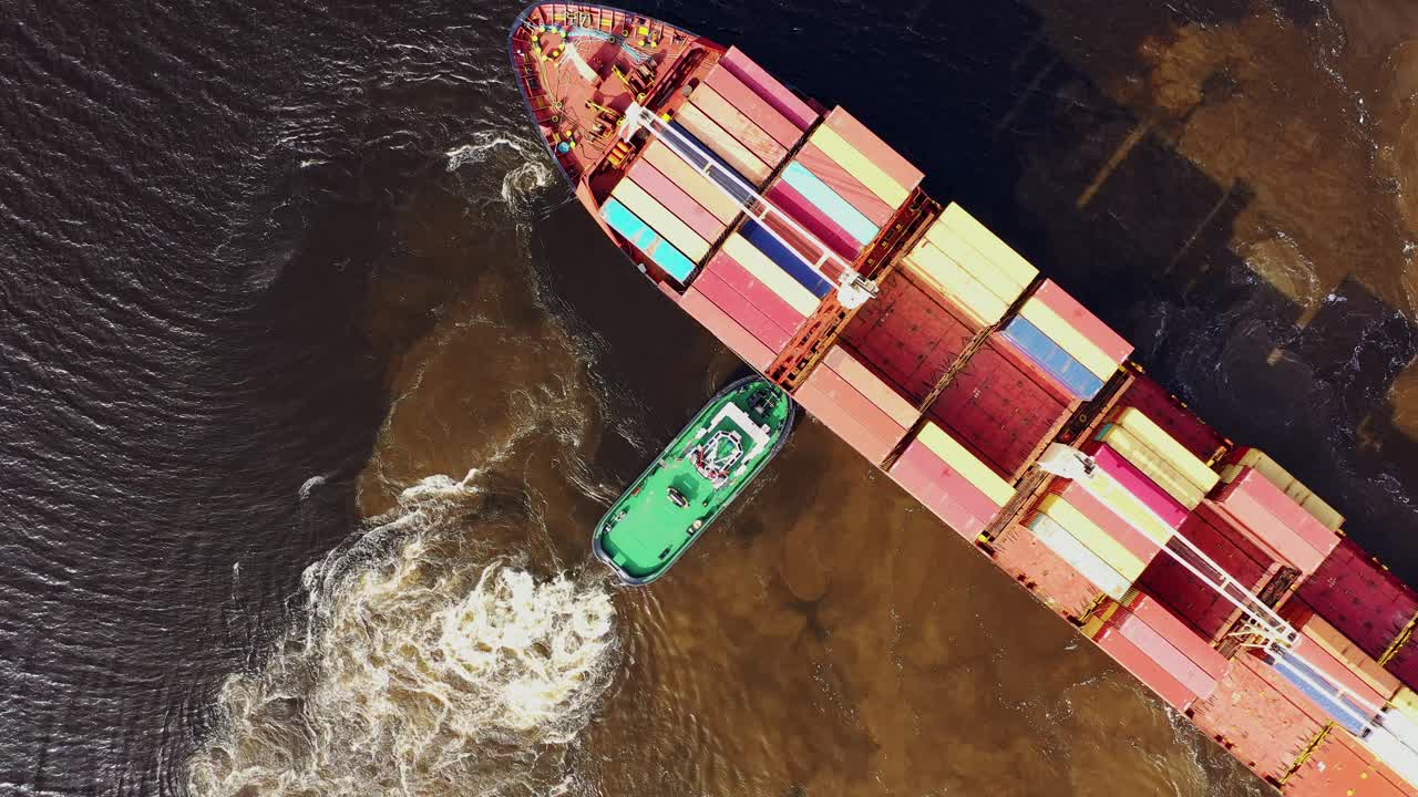 Drone kredser nedad, mens slæbebåd styrer containerskib gennem mudret flodvand