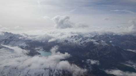 High-angle-drone-shot-above-clouds-during-sunny-day-showing-snowy-summits-of-mountains-in-Austria---Blue-lake-in-the-valley