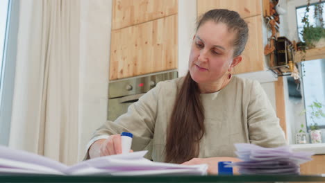 close-up view of woman applying liquid glue with glue stick to paper on table, working on paper craft in well-lit home kitchen environment with papers stacked nearby
