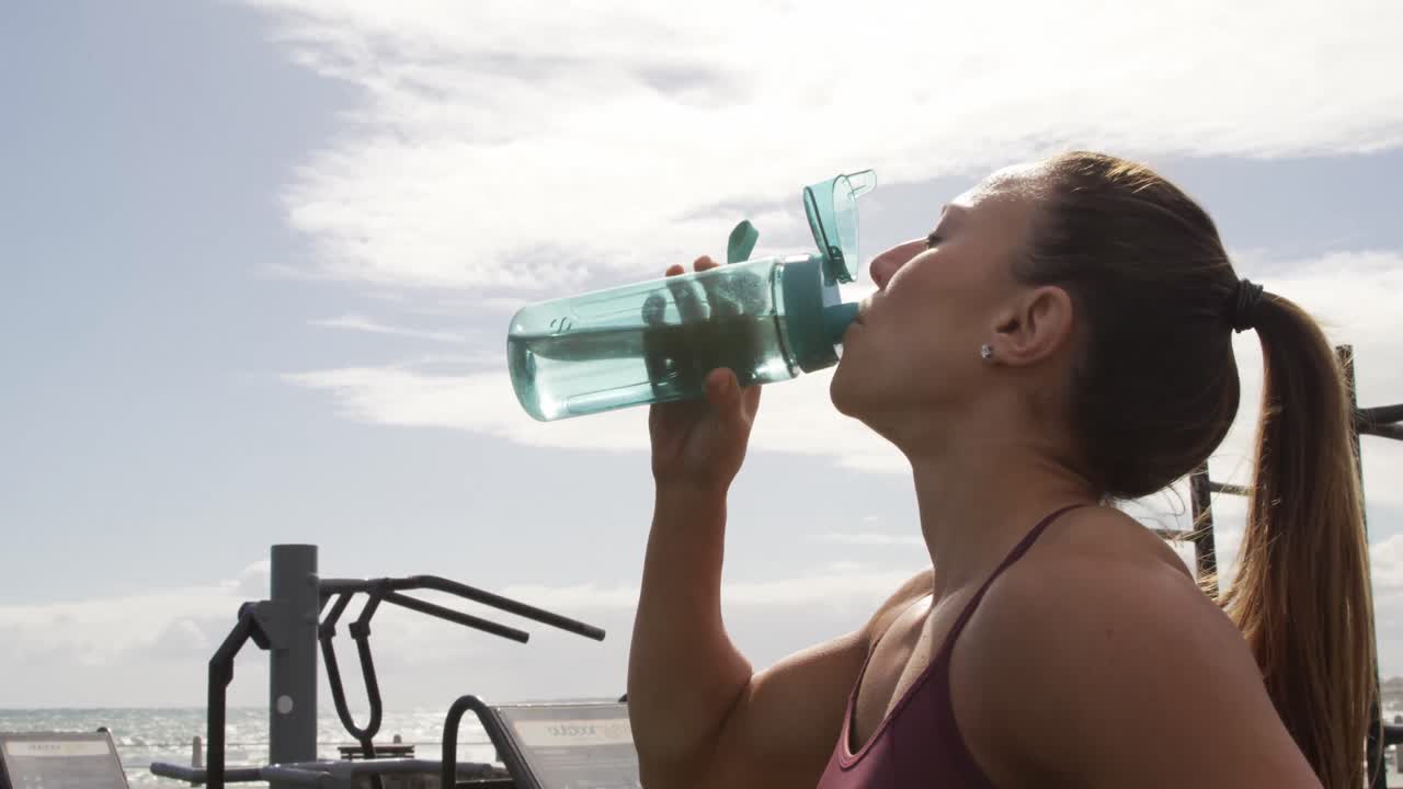 mujer caucásica deportiva bebiendo agua en un gimnasio al aire libre durante el día