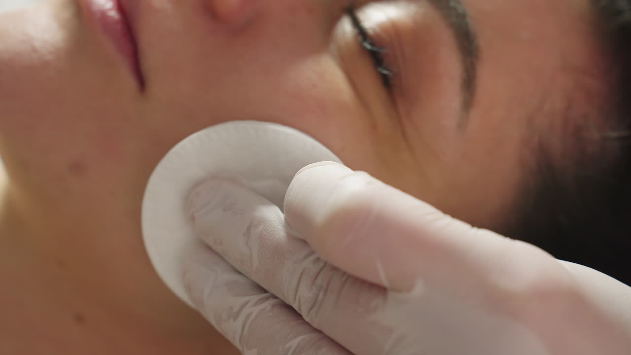 Close up of young woman face as gloved therapist hand uses soft wool pad to rub from cheek to forehead during soothing spa facial treatment under soft ambient lighting in bright clinic environment