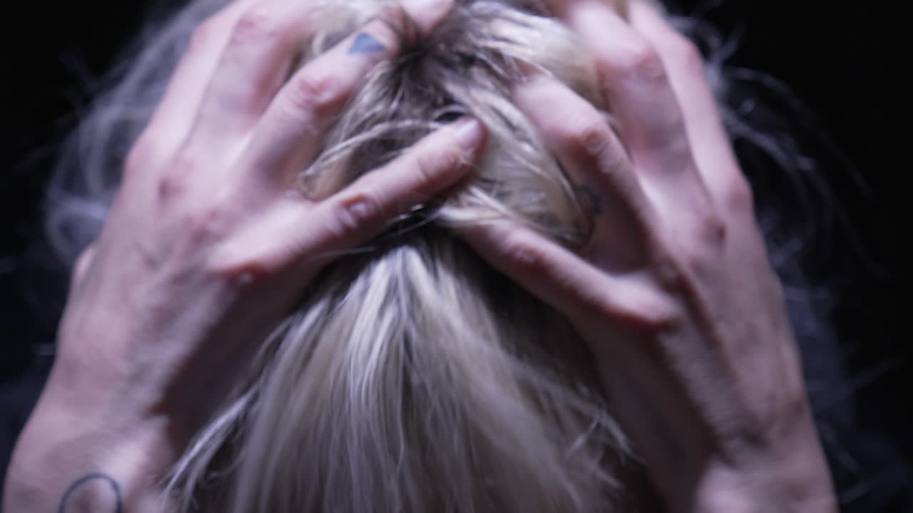 금발의 우울한 여자가 그녀의 머리를 잡고, 머리를 긁고, 필사적으로 머리카락을 잡아당기고, 머리 위 조명 하나가 비추는 희미한 방에서