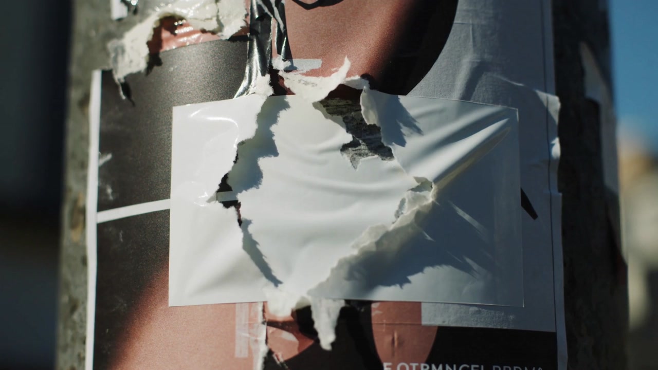 Lifting white adhesive sheet by breeze and revealing torn poster torso on metal street pole, tape