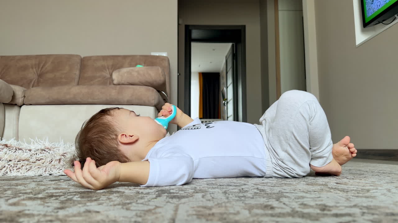 Child in white clothes lies on the floor holding a pacifier. Peaceful baby boy watches TV in the living room.