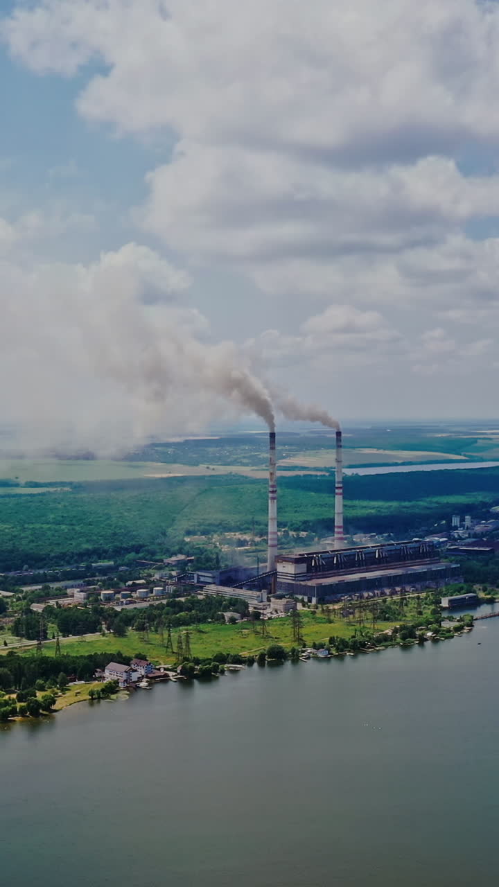 Aerial view of plant on the bank of the river in summer. View from above on natural background of beautiful river and factory with smoke from pipes. Slow motion. Camera moves up. Vertical video