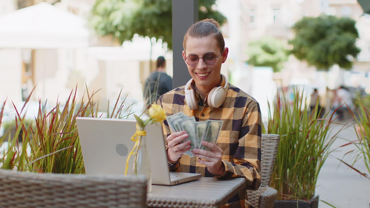 Happy rich man use laptop winner holding fan of cash money in dollar banknotes victory in city cafe