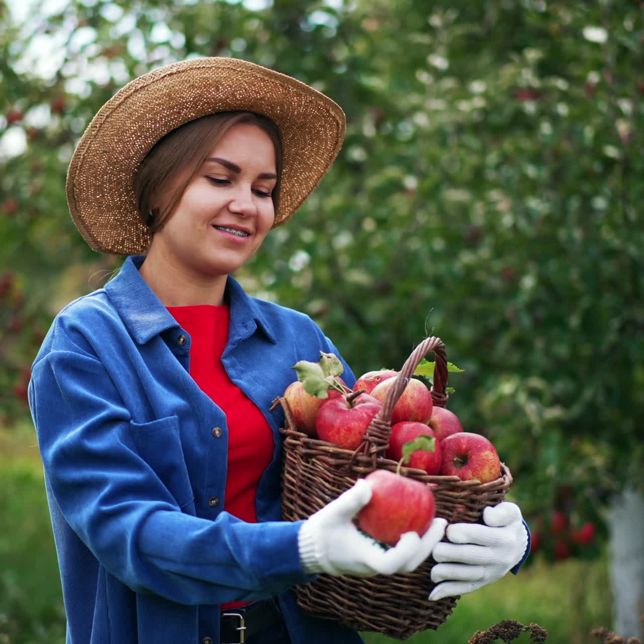 Smiling lady in blue shirt and straw hat holding a basket full of apples. Female farmer takes a fruit and tosses it in the air. Nature backdrop