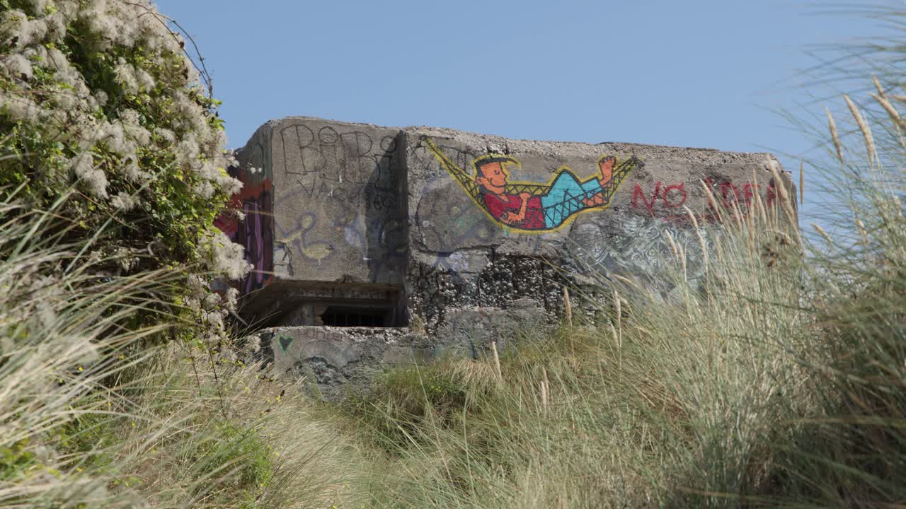 Static wide shot of abandoned concrete bunker with colorful street art, bright daylight, grassy dunes