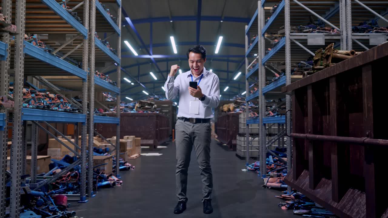 Figura intera di un lavoratore professionista maschio asiatico in piedi con il suo smartphone, in un magazzino industriale di rottami metallici e riciclaggio. Alza il pugno con un urlo di gioia.