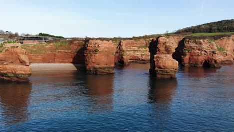 panoramic aerial drone view of the clear and calming waters with a majestic, grass-covered rock pinnacles along the cliffside of ladram bay