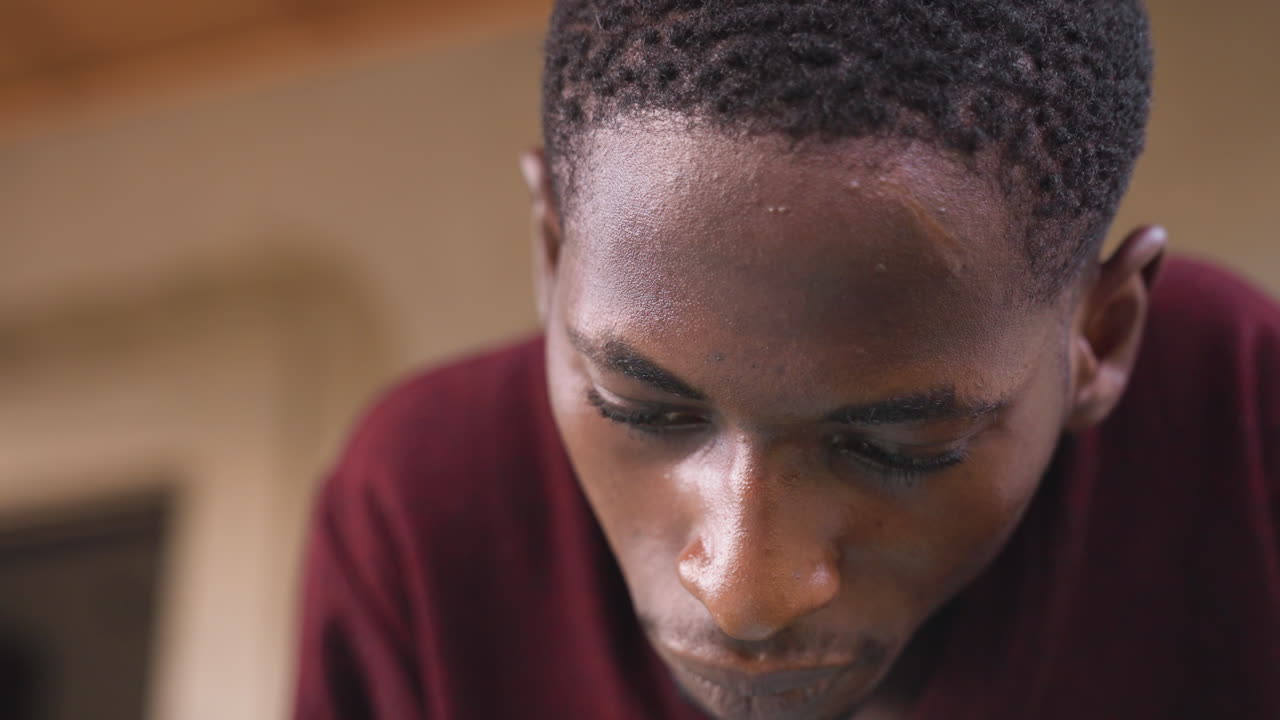 Close-up de jovem inclinado para a frente, suor brilhando na testa, camisa bordô capturando luz quente, fundo de varanda desfocado, olhar introspectivo para baixo, atmosfera íntima e melancólica mostrando textura.