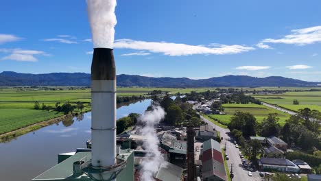 drone footage captures a factory's smoke stack releasing steam over lush green fields and a river under a clear sky