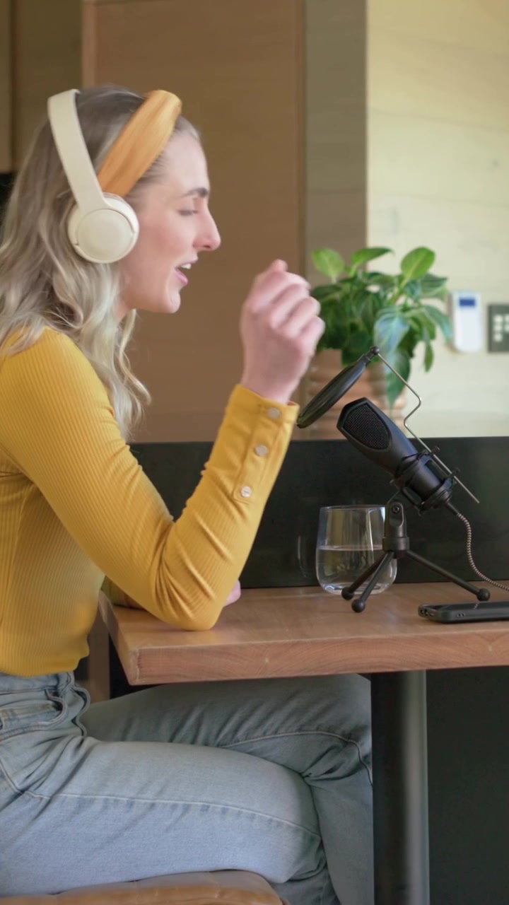 Vertikal video: En kvinde i sennepsgul top læner sig mod mikrofonen og taler i et køkkenhjørne, mens hun justerer sit headset.
