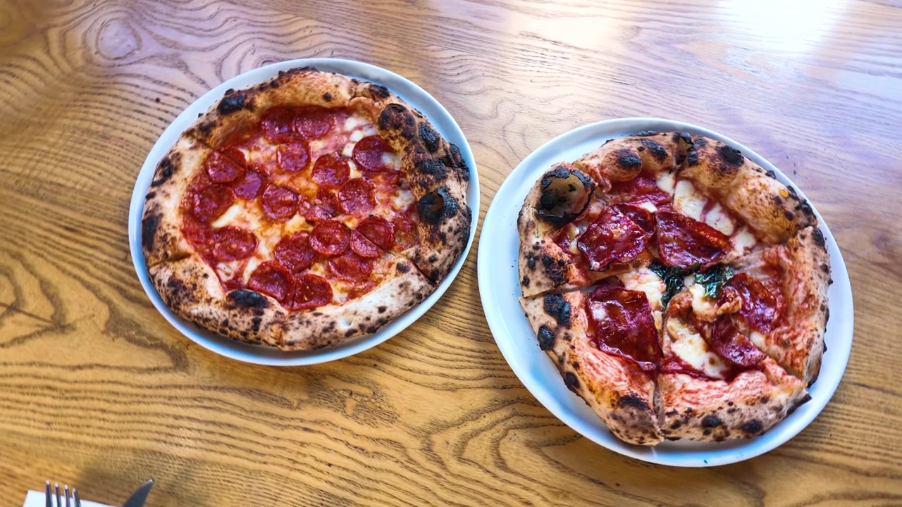 Fugleperspektiv af pepperoni- og prosciuttopizzaer på et træbord i naturligt lys.
