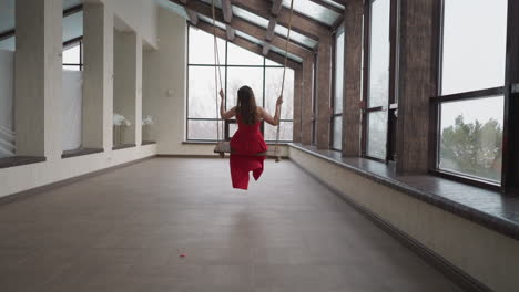 woman in crimson dress swings on closed terrace. dreaming lady wearing stylish gown with waving skirt plays swings in residence. glamour and fashion