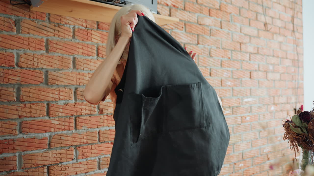 Decorator with blonde hair in orange shirt wearing black apron stands and takes off the apron against rustic brick wall, with dried flower decorations in glass vases, preparation and design