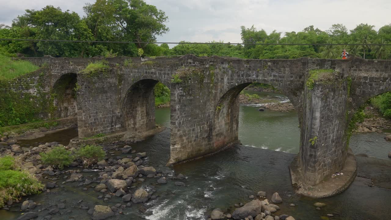 A wide shot of a historic stone bridge with multiple arches spans shallow rocky river in Tayabas, Quezon Province, Philippines, surrounded by lush green trees and serene rural landscape