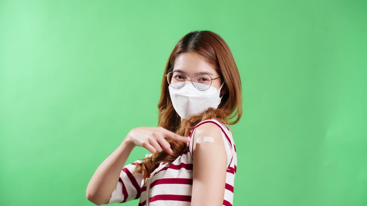 The portrait of an Asian woman with eyeglasses is smiling on a mask being positive emotions and Cheerful vaccinated showing arm with medical patch and laughs plaster on her shoulder