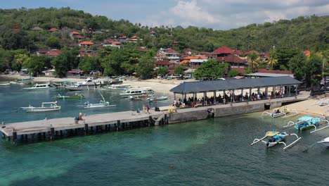 tourists-on-a-pier-at-padangbai-port-waiting-for-a-ferry-from-Bali-Indonesia-with-white-sand-beach-and-fishing-boats-anchored-in-turquoise-blue-water,-aerial