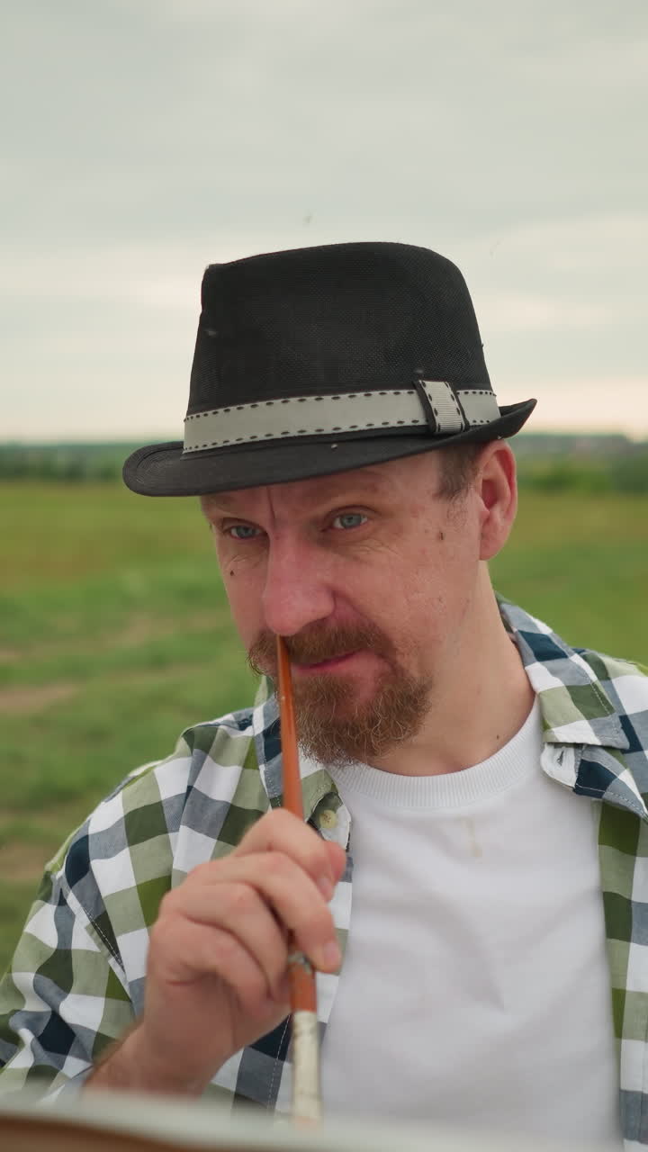 모자와 체크색 셔츠를 입은 화가의 클로즈업, 잔디에 서서 페인트 브러쉬로 코를 는 모습. 배경은 구름이 가득한 하늘 아래의 시골 풍경을 보여줍니다.