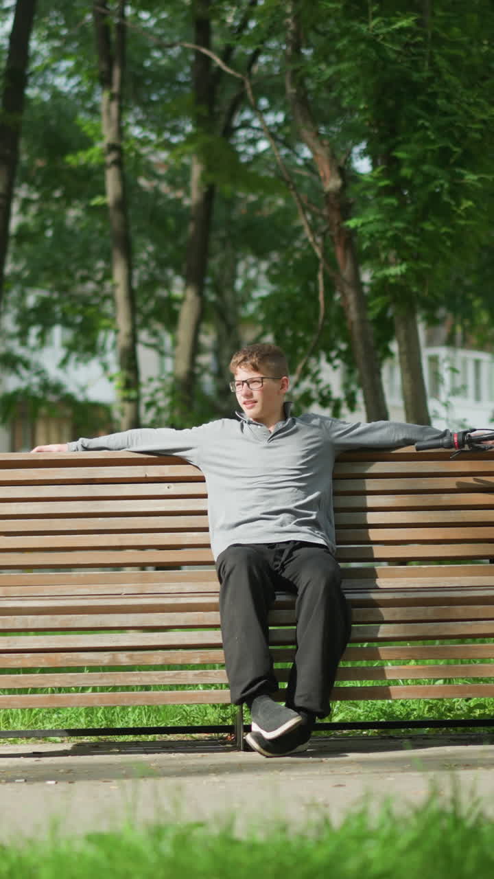 안경을 쓴 소년, 회색 은 공원에서 나무 벤치에 앉아, 자신을 조정하는 동안 앞으로 어, 왼쪽 다리에 오른쪽 발을 놓고, 근처에 주차 된 자전거, 초록색과 나무 주변