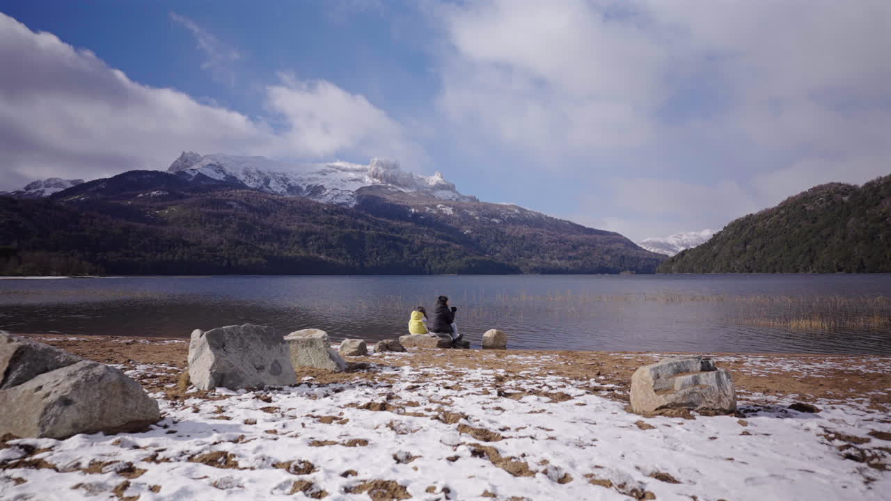 Woman and child sit by the snowy shore of Lago Falkner in Neuquén, Argentina, drinking mate and admiring the serene, mountain-framed lake, static camera, real time