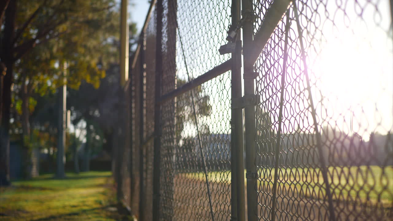 levantándose a lo largo de una puerta de valla de eslabones de cadena con cerraduras al amanecer fuera de un campo de béisbol de hierba en un parque público