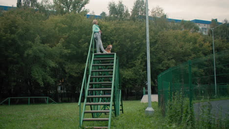 female pet therapist resting on high ladder trainer with dog during break from training activity in lush green park surrounded by trees near tennis court under calm cloudy sky reflecting bond