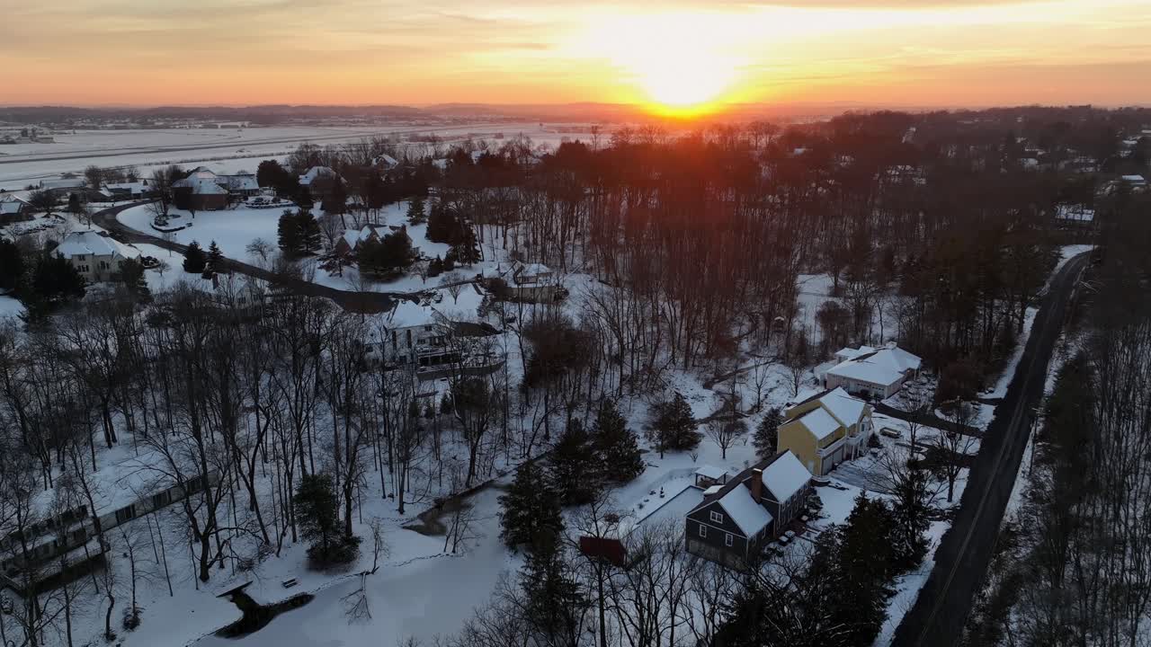 Talvinen auringonlasku lumisten talojen, puiden ja kumpuilevien kukkuloiden yllä rauhallisessa lähiössä, lämmin iltavalo. Tyyni ja rauhallinen tunnelma Yhdysvalloissa. Panoraamanäkymä ilmasta. PA, US