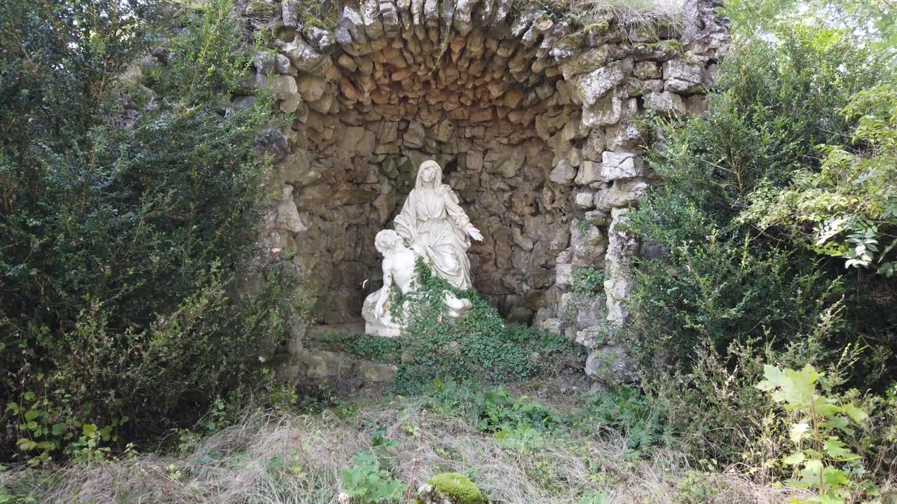 A serene view of Sion Hill in Vaudémont, featuring a stone grotto with a religious statue, surrounded by lush greenery and trees.