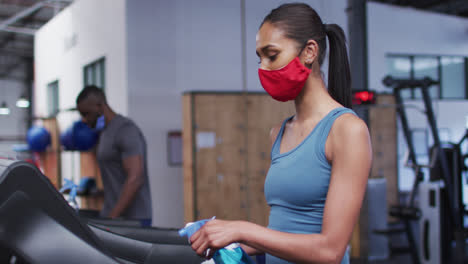 Mujer-Caucásica-En-Forma-Con-Mascarilla-Limpiando-La-Cinta-De-Correr-Con-Desinfectante-En-El-Gimnasio