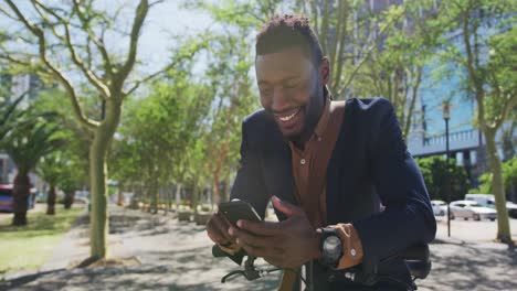 smiling african american businessman using smartphone leaning on bike