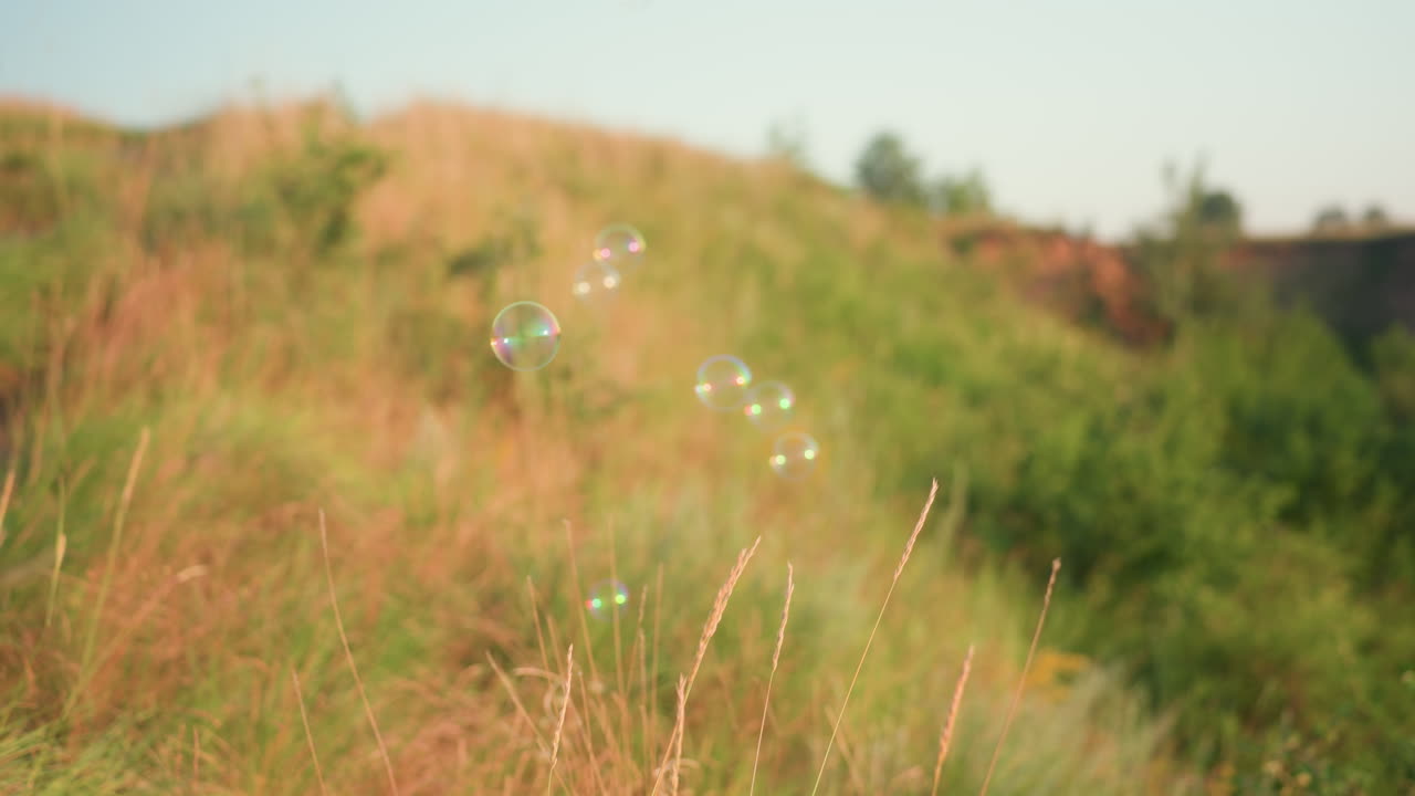 Iridescent soap bubbles drifting over sunlit grass hillside under pastel sky catching warm golden hour rays as they float gently into distance against serene nature backdrop