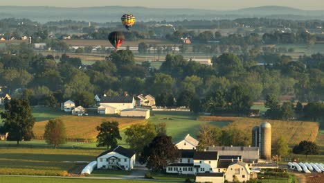 Drone-wide-shot-of-american-countryside-with-farm-houses-and-flying-hot-air-balloon-in-the-air-at-sunset-time