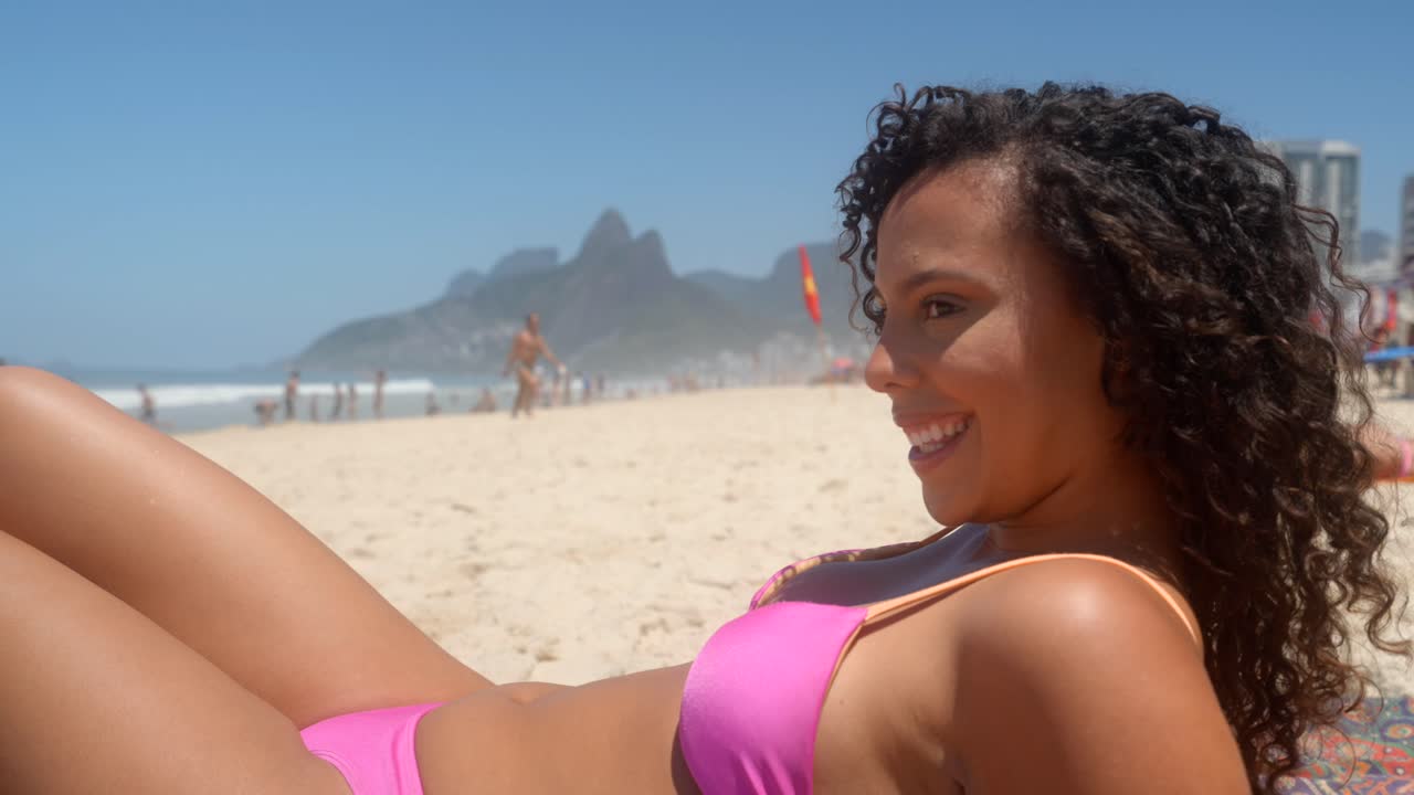 A beautiful multiracial woman in a pink bikini lies on her back, smiling and enjoying the sun on Ipanema beach with the Two Brothers Mountain in the background