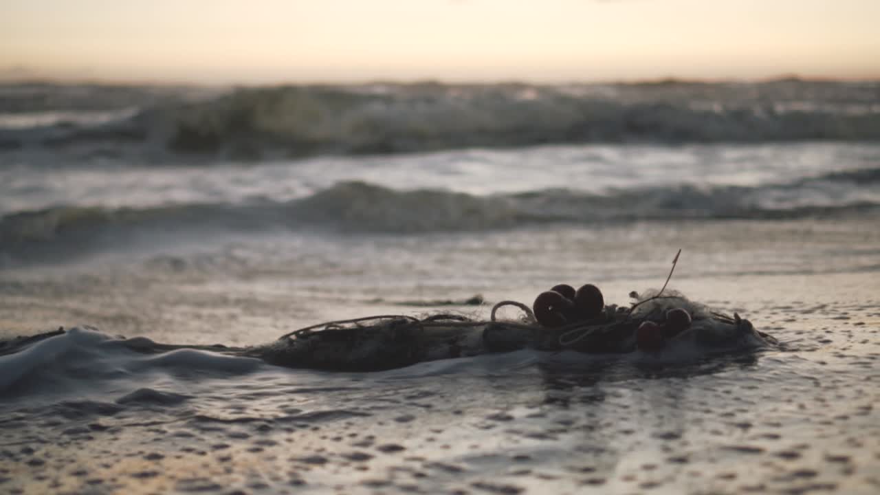 SLOW MOTION of a fisherman's net left abandoned on the shoreline and bathed by sea water during the golden hour with waves in a blurry background