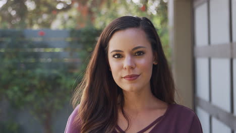 Portrait-Of-Smiling-Young-Hispanic-Woman-In-Garden-At-Home-Against-Flaring-Sun