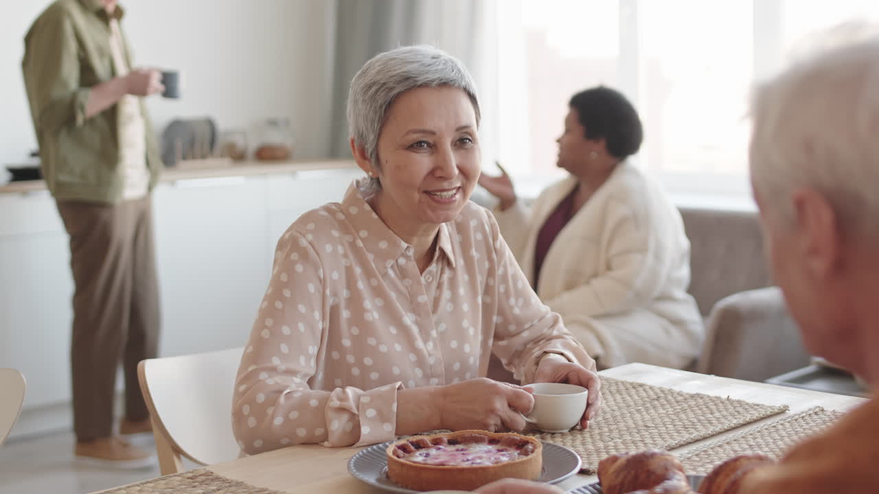Waist-up over shoulder of joyful senior Asian woman smiling, sitting at table, having tea, talking to unrecognizable friend