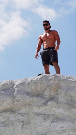 shirtless bodybuilder with sunglasses and cap on the hill in a sunny day. strong athlete with muscular body under blue sky outdoors. vertical video