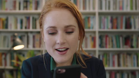 portrait of beautiful young red head business woman intern texting browsing using smartphone looking surprised excited reading text messages in bookshelf background