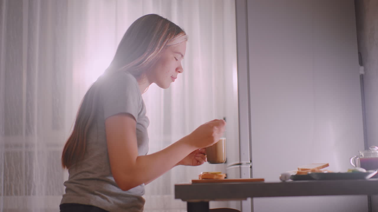 Ragazza che mescola il caffè al tavolo della cucina con un panino e del pane accanto, mentre si gode un tranquillo momento di colazione con la luce del sole che filtra attraverso la tenda creando una calda e serena mattinata