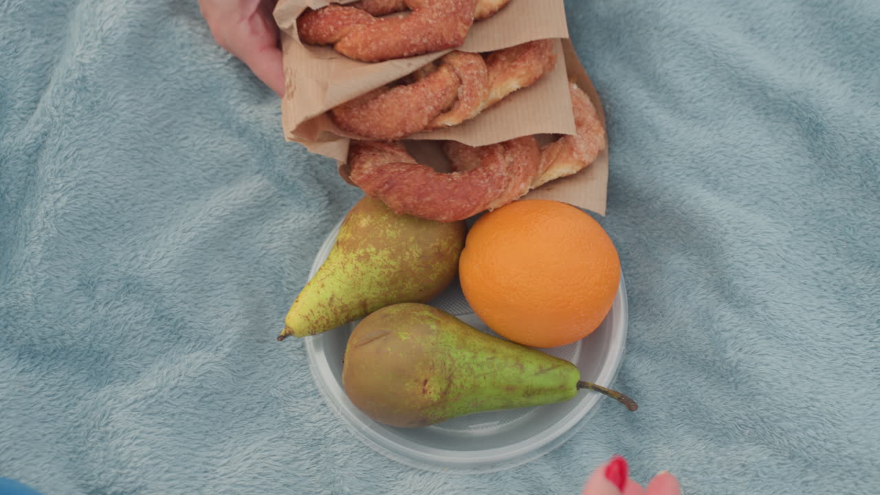 mature women hand view placing baked bread and plate of orange on picnic mat, soft towel on grass, summer park setting, preparing snack, sharing moment, warm light, friendship and relaxation outdoors