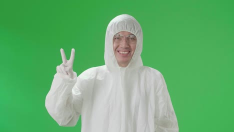 asian male wearing personal protective equipment uniform ppe smiling to camera with a victory hand sign in green screen studio