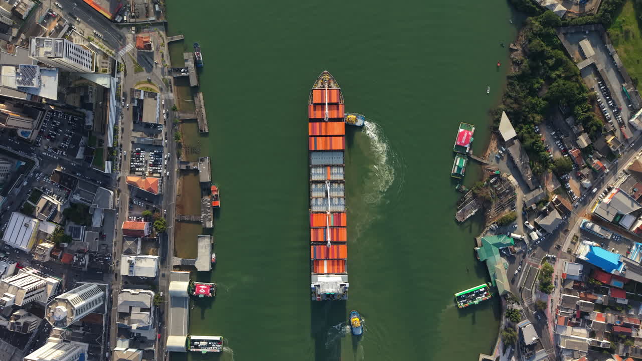 Container ship moves through the Itajaí Port channel in Santa Catarina, Brazil, guided by tugboats between urban docks and industrial areas, captured from above in a precise top-down slow drone shot