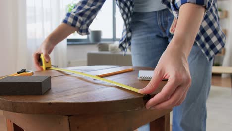 Woman-with-Ruler-Measuring-Table-for-Renovation.repair,-diy-and-home-improvement-concept--woman-with-ruler-measuring-old-round-wooden-table-for-renovation-and-writing-to-notebook