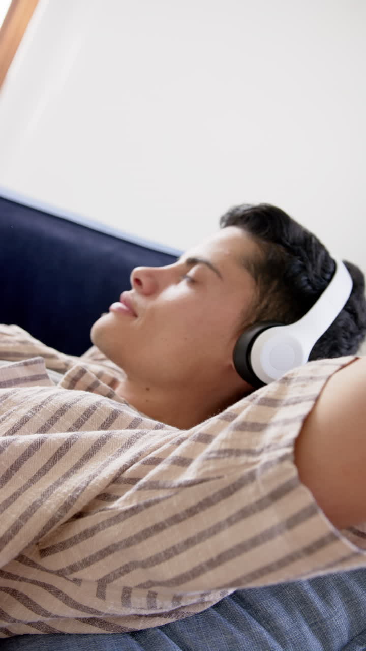 Vertical video of biracial man in headphones lying on couch, slow motion