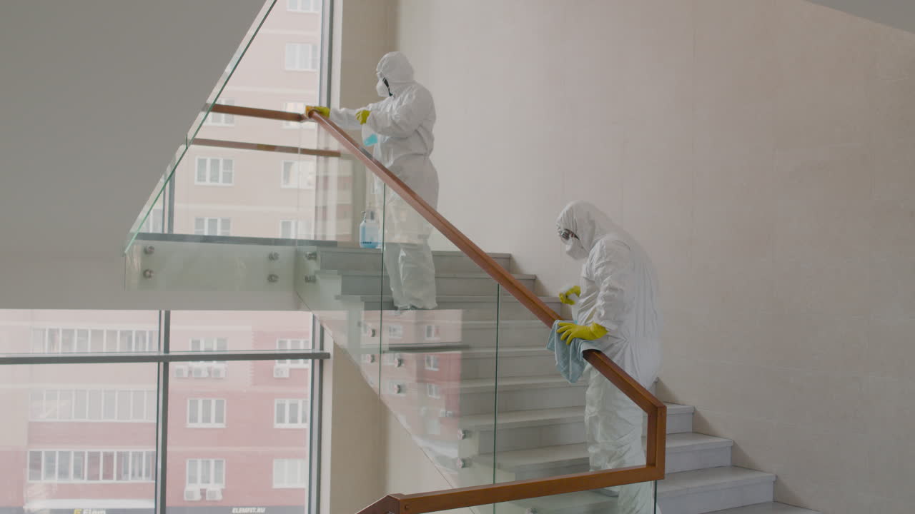 vista trasera de dos hombres de limpieza que usan equipo de protección personal limpiando barandillas de escaleras dentro de un edificio de oficinas