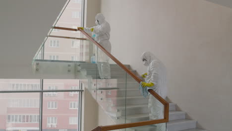 rear view of two cleaning men wearing personal protective equipment cleaning stair railings inside an office building